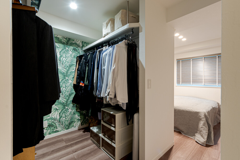 ご夫婦の寝室横に新設した大容量のW.I.C。扉を設置しないことで、スムーズな動線と共に解放感も感じることができるようになりました。もちろん、収納内の枕棚の上には、普段使わない季節物をたっぷり収納することができます。リゾート地「ハワイ」をイメージするモンステラ柄のアクセントクロスもポイントです。